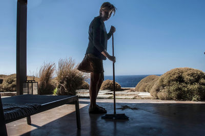 Woman cleaning porch with broom against blue sky