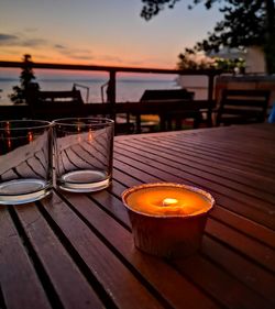 Lit tea light candle by empty glasses on table against sky during sunset