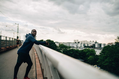 Portrait of tired sportsman leaning on railing at footbridge against sky