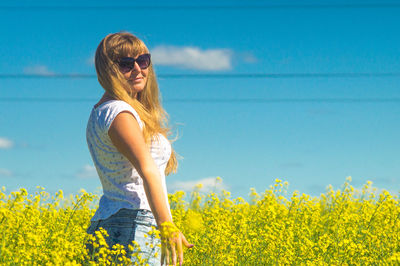Side view of young woman standing amidst yellow flowering plants on field against sky during sunny day