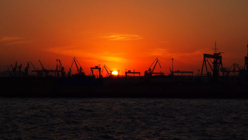 Silhouette cranes at commercial dock against orange sky