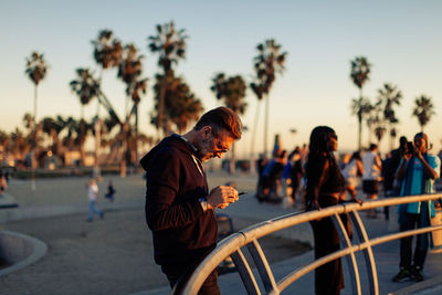 Adult man with beard looking at his phone during sunset at a skatepark with palm trees in background 