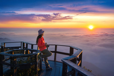 Rear view of woman standing by railing against sky during sunset