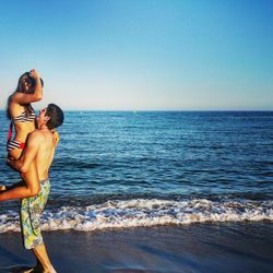 Shirtless man lifting girlfriend on shore at beach against clear blue sky