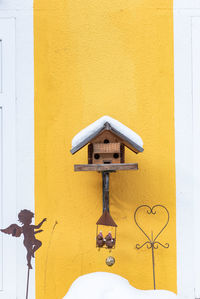 Homemade wooden bird's feeder in winter, under snow. yellow and white wall as a background.