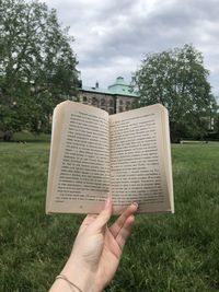 Cropped hand holding book on field against sky