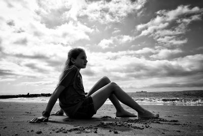 Side view of girl sitting at beach against cloudy sky