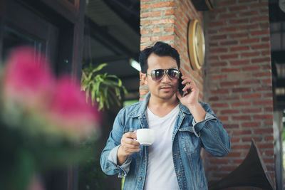 Young man holding coffee cup while using smart phone against brick wall at cafe