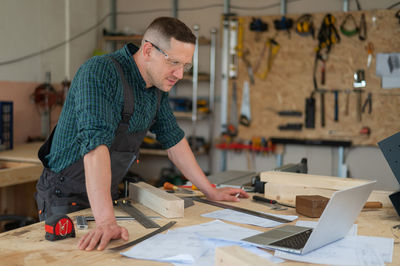 Portrait of a carpenter in protective glasses and work overalls uses a laptop in a workshop