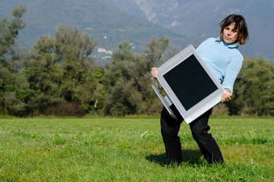 Portrait of woman carrying heavy television set on grassy field