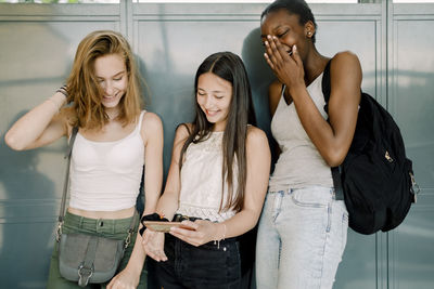 Smiling junior high students with friend using mobile phone while standing in school corridor