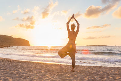 Full length of woman meditating on beach against sky during sunset