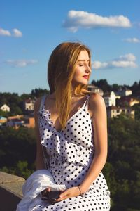 Beautiful young woman sitting on retaining wall against sky