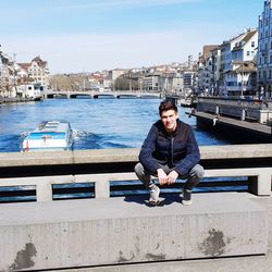 Portrait of man sitting on railing by canal against sky