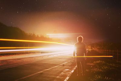 Rear view full length of man standing by light trails at roadside during night