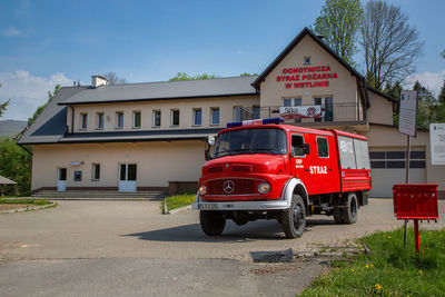 Red car on street by buildings against sky