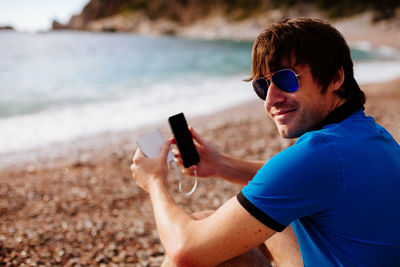 Portrait of man holding smart phone while sitting at beach