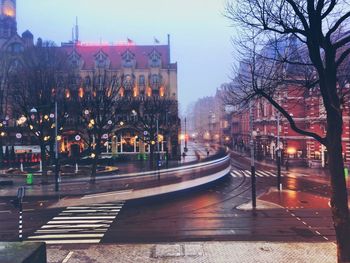 Illuminated street by buildings against sky at dusk