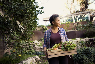 Thoughtful woman carrying crate full of freshly harvested vegetables at organic farm