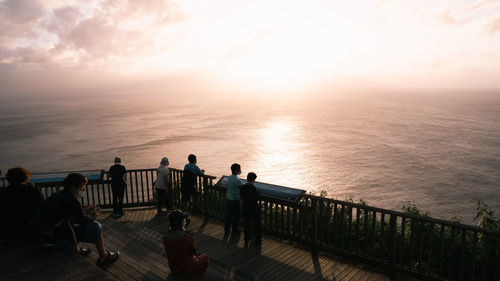 People sitting on railing by sea against sky during sunset