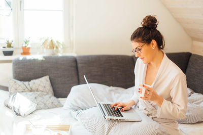 Young woman holding coffee cup using laptop while sitting on bed at home