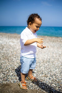 Happy kid in white shirt and blue shorts plays with pebble on the beach