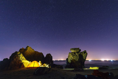Illuminated tent by rock formations against sky at star field