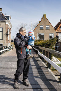 Full length of grandfather carrying grandchild while standing on boardwalk in city