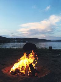 Campfire burning at lakeshore against cloudy sky