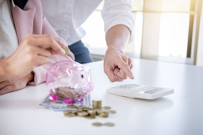 Midsection of woman with colleague putting coin in piggy bank at table
