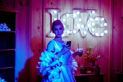 Portrait of young woman in dress standing against illuminated wall