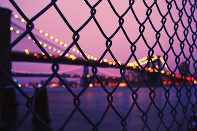 Illuminated bridge over river seen through silhouette chainlink fence against clear sky during sunset