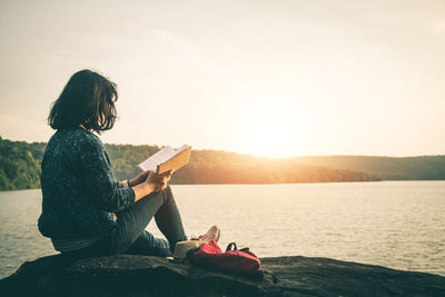 Man sitting on book by sea against sky during sunset