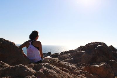 Rear view of woman sitting on rocks by sea against clear sky