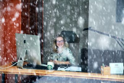 Portrait of young woman using computer seen through window