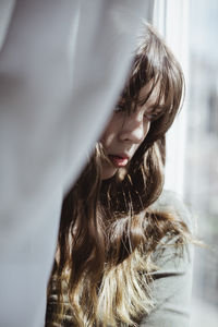 Close-up of young woman hiding behind window curtain