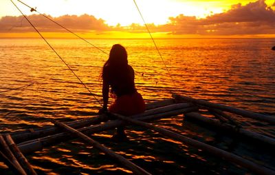 Rear view of silhouette person sitting on sea against sky during sunset