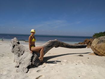 Portrait of mid adult woman in bikini sitting on log at beach against sky during sunny day