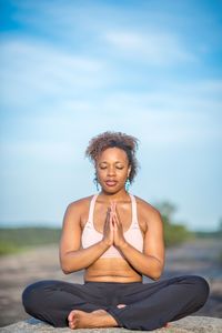 Mid adult woman meditating while sitting on rock against blue sky