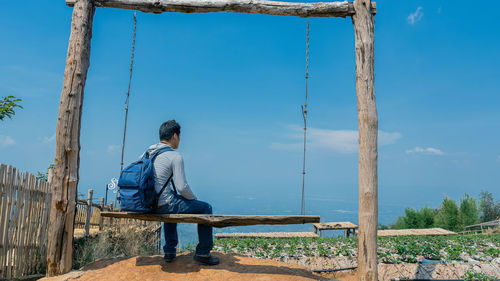 Rear view of backpacker sitting on swing against sky