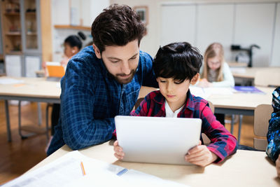 Teacher assisting male student in using digital tablet while sitting in classroom