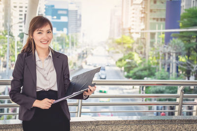 Portrait of young woman holding file standing by railing