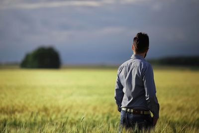 Rear view of man standing amidst wheat field against cloudy sky