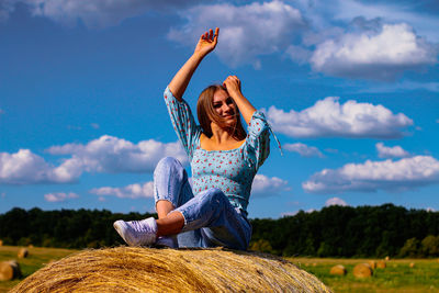 A young girl sits on a bale of hay with her arms raised and smiles