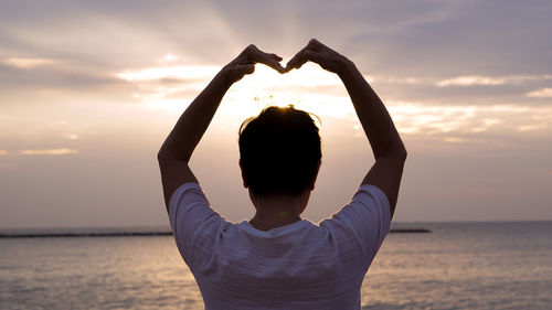 Rear view of woman making heart shape while standing by sea against sky