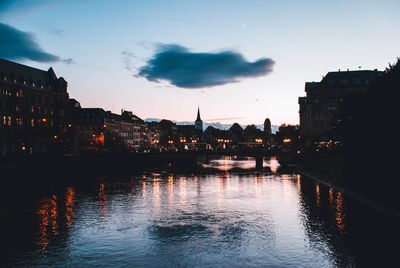 Illuminated buildings by river against sky at sunset