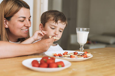 Cute toddler boy with his mother having breakfast with puncakes and berries in kitchen at home