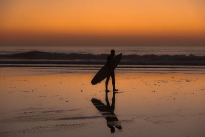 Silhouette woman walking on beach against orange sky