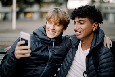 Smiling teenage boy taking selfie through smart phone while sitting with friend in city