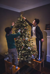 Two boys standing on chairs to decorate the top of a christmas tree.